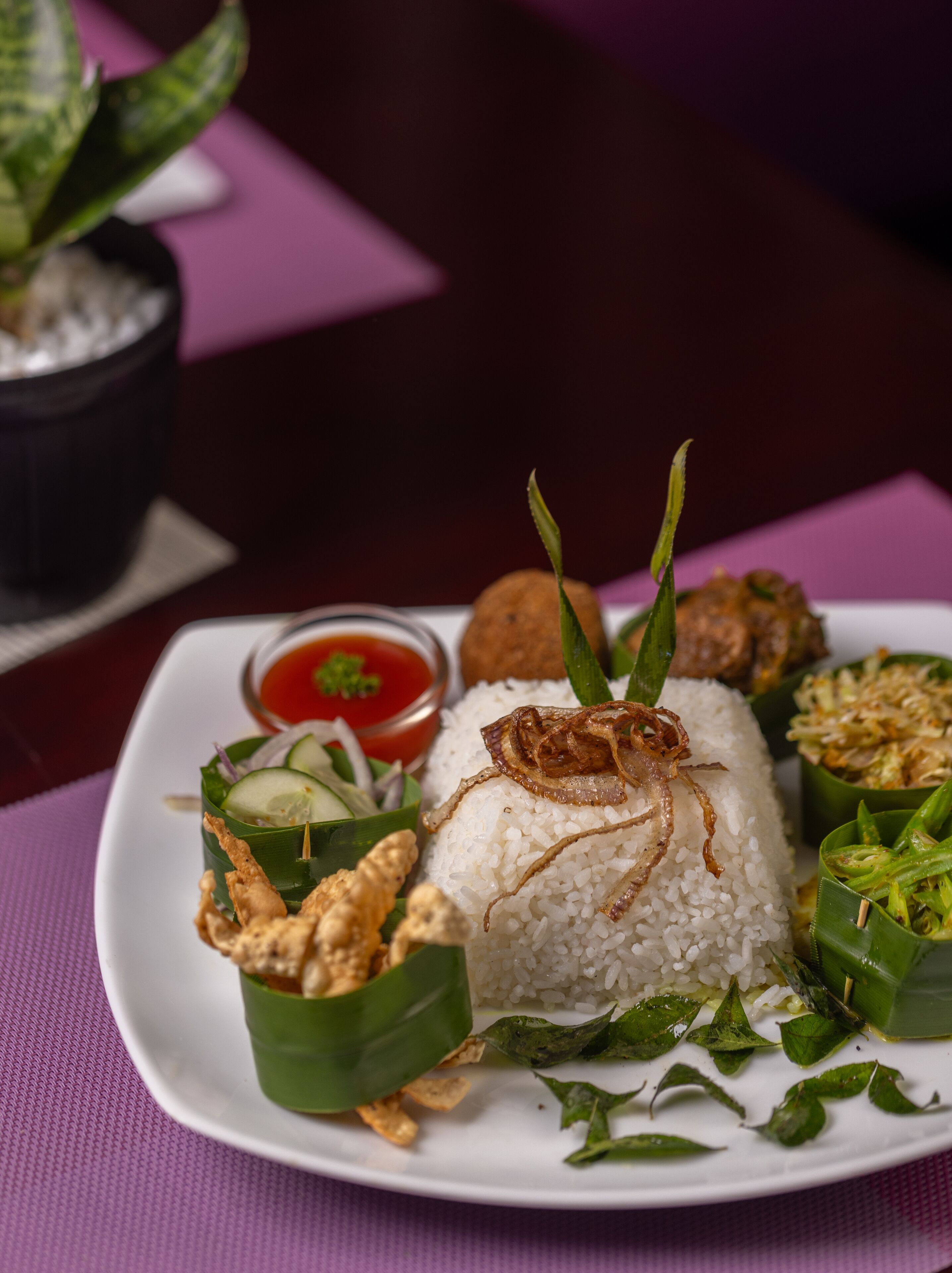 Une assiette de riz blanc garnie d’oignons frits, accompagnée d’un petit bol de sauce rouge, d’un morceau de poulet frit et d’une salade de concombres.