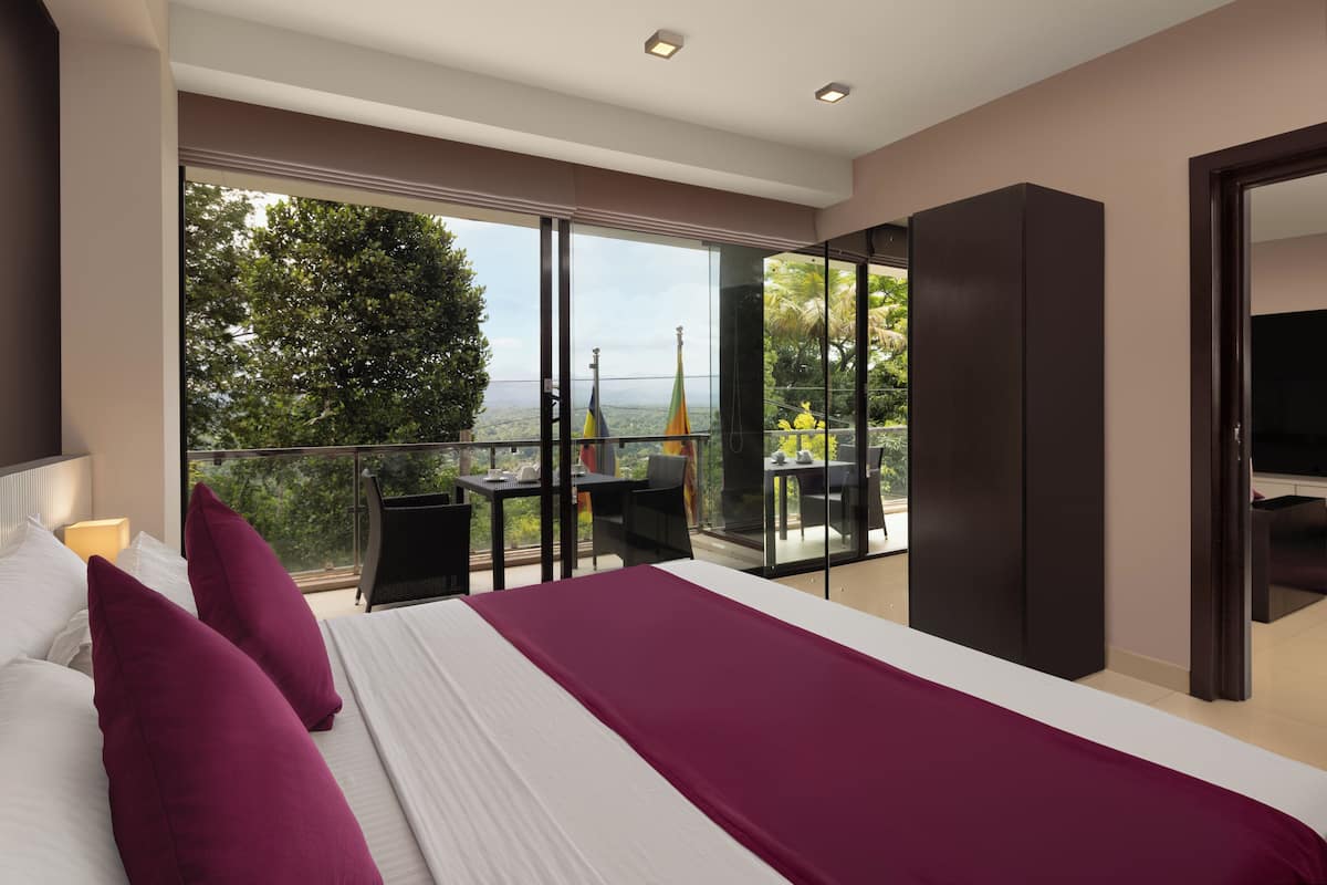 A modern hotel room with a large bed, a balcony with a table and chairs, and a view of the surrounding landscape.