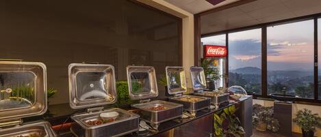 A buffet area with multiple food dispensers, a Coca-Cola sign, and a view of the cityscape through large windows.