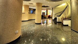 Un vestíbulo de hotel con suelos de mármol, amplias columnas, una recepción y una escalera.