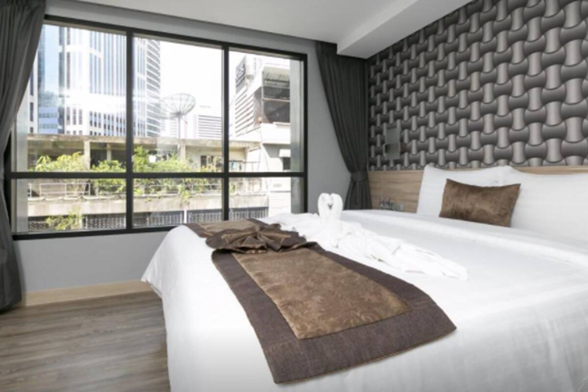 A modern hotel room with a large bed, a view of buildings through the window, and a textured wall behind the bed.