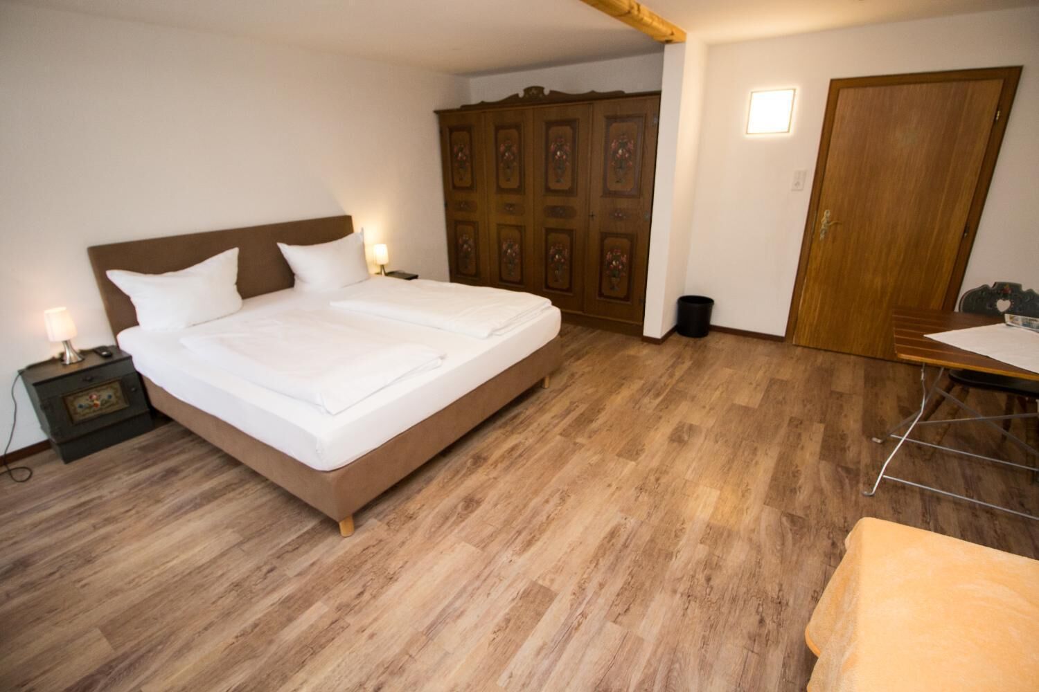 A bedroom with a large bed, wooden wardrobe, and a small table with a chair.