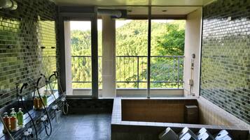 A bathroom with a bathtub, a window with a view of greenery, and a tiled wall with a faucet and soap dispenser.