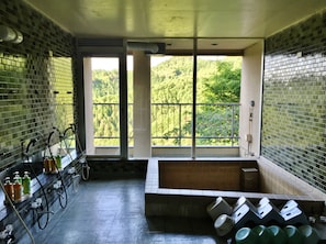 A bathroom with a bathtub, a window with a view of greenery, and a tiled wall with a faucet and soap dispenser. - Ebisukan (Yoshino)