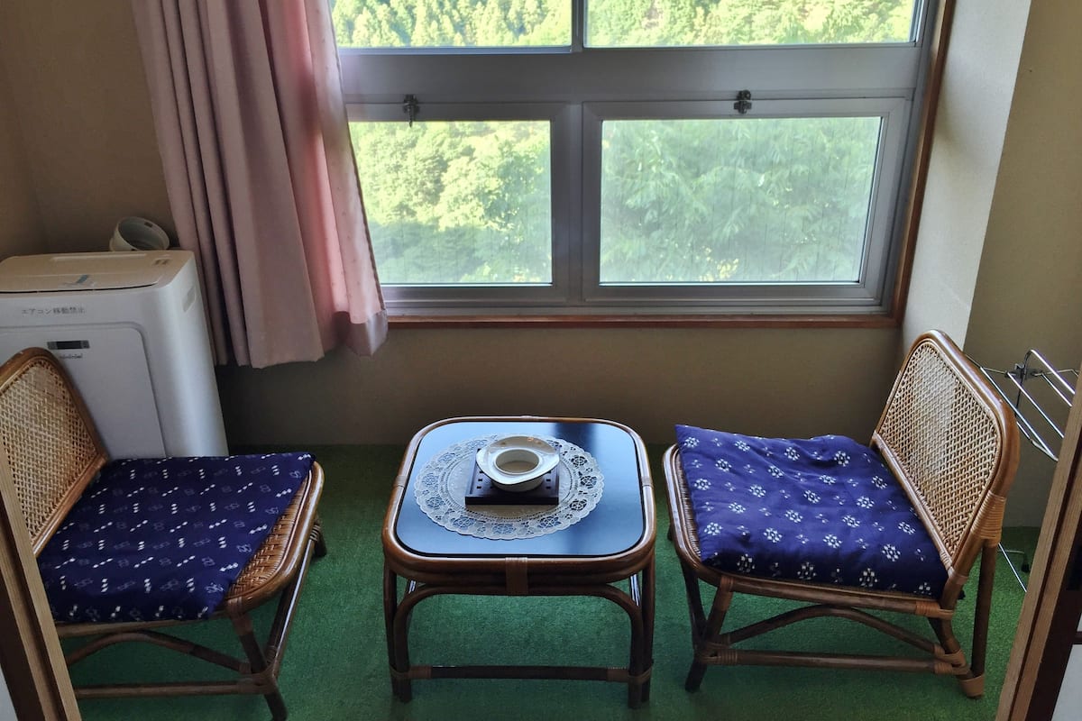A room with two wicker chairs, a small table with a lace tablecloth, and a window with a view of a forest.