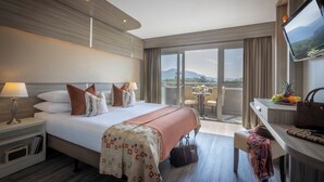 Une chambre d’hôtel avec un grand lit, un bureau avec une télévision, un balcon avec vue et un panier de fruits.