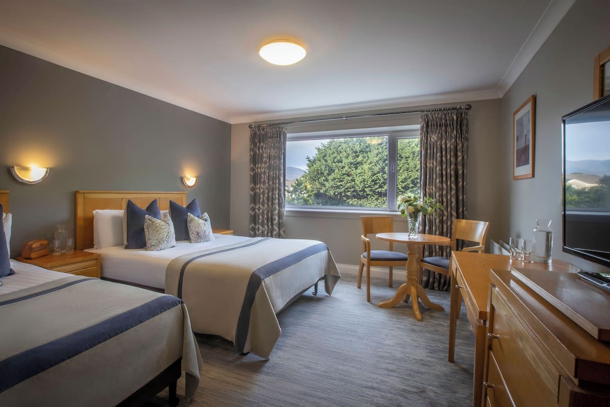 Ein Hotelzimmer mit zwei Betten, einem kleinen Tisch mit Stühlen, einem Fernseher und einem Fenster mit Vorhängen.