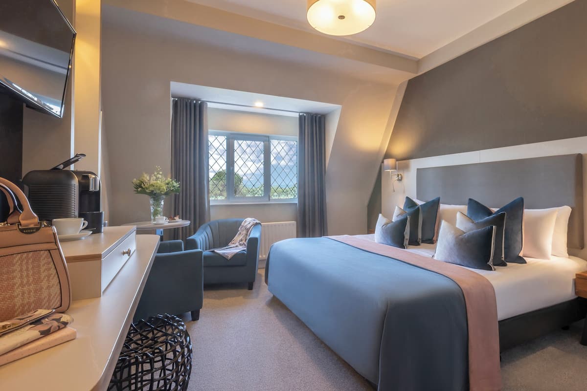 Ein modernes Hotelzimmer mit einem großen Bett, einem Schreibtisch mit Stuhl, einer Kaffeemaschine und Blick ins Freie durch ein Fenster.