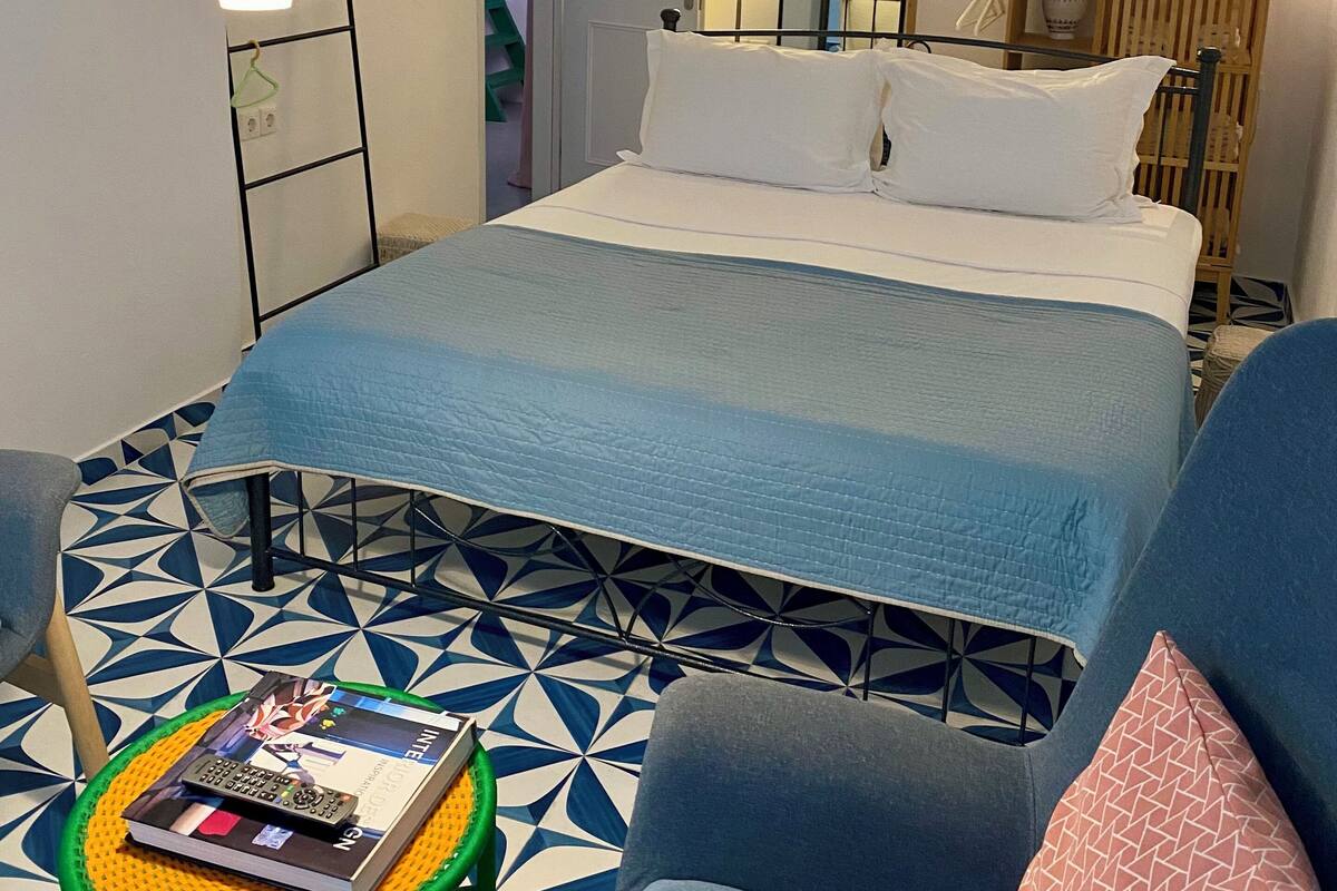 Un dormitorio con cama, una silla, una mesita con libros, una lámpara colgante, un espejo y un aire acondicionado instalado en la pared.