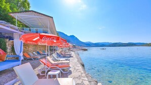 A poolside area with lounge chairs and red Coca-Cola umbrellas, a stone wall, and a view of the sea and mountains. - Olympos Mocamp Kas (Kas)