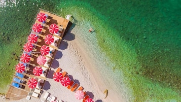 Vista aérea de un resort frente a la playa, con una terraza de madera, varios sombrillas rojos y blancos, y agua turquesa cristalina.