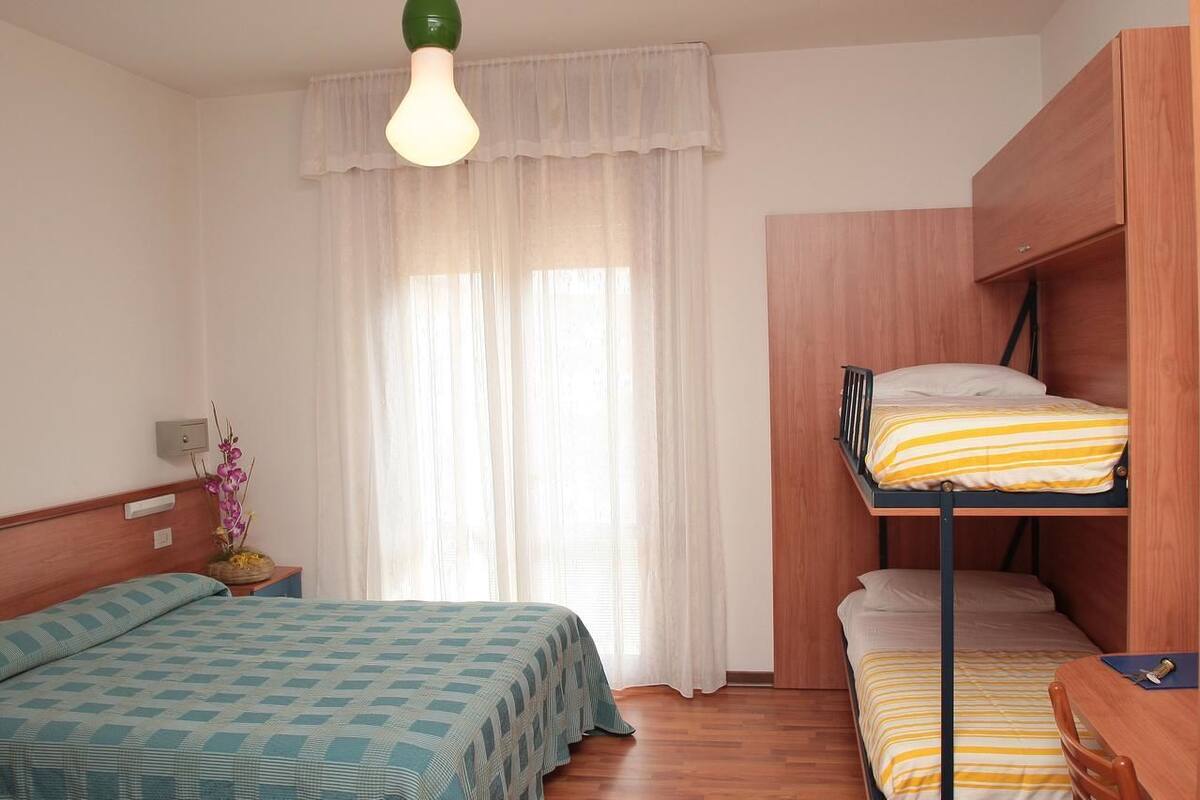 A room with a bunk bed, a single bed, a bedside table with a lamp, and a window with sheer curtains.