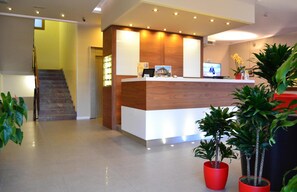 A reception area with a wooden reception desk, a TV screen, and potted plants. - Hotel Midi (Jesolo)