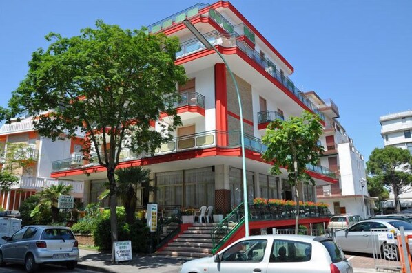 A multi-story hotel with a red and white facade, balconies, and a staircase leading to the entrance. - Hotel Midi (Jesolo)