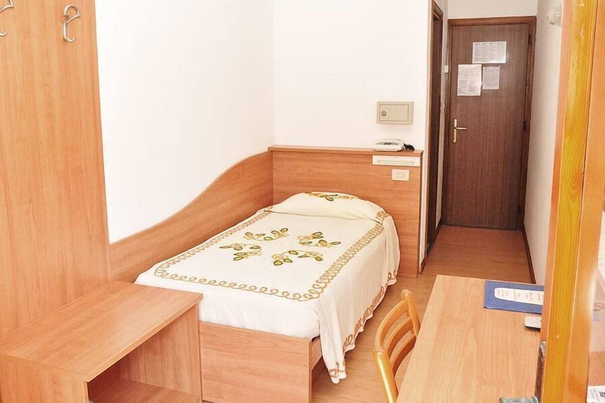 A small, neatly arranged bedroom with a single bed, a bedside table, a chair, and a wooden wardrobe.
