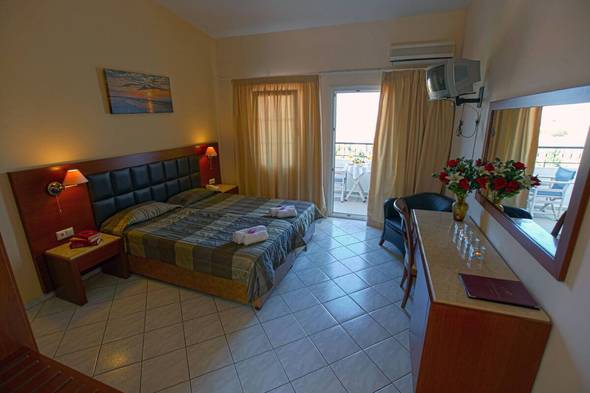 Une chambre d’hôtel avec un lit, des lampes de chevet, une petite table avec un livre, un vase de fleurs et un balcon avec une table et des chaises.