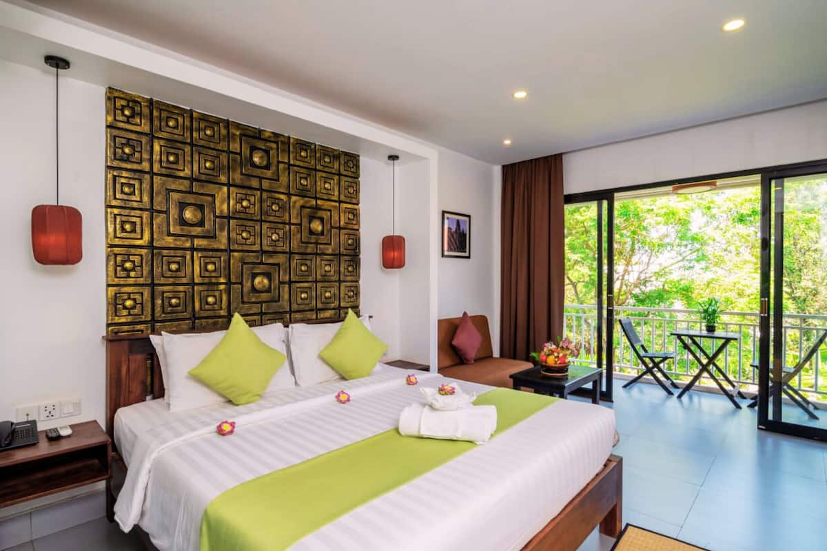 Habitación de hotel con una cama grande, un panel decorativo en la pared, una mesita con una planta, una silla y un balcón con vistas a zonas verdes.