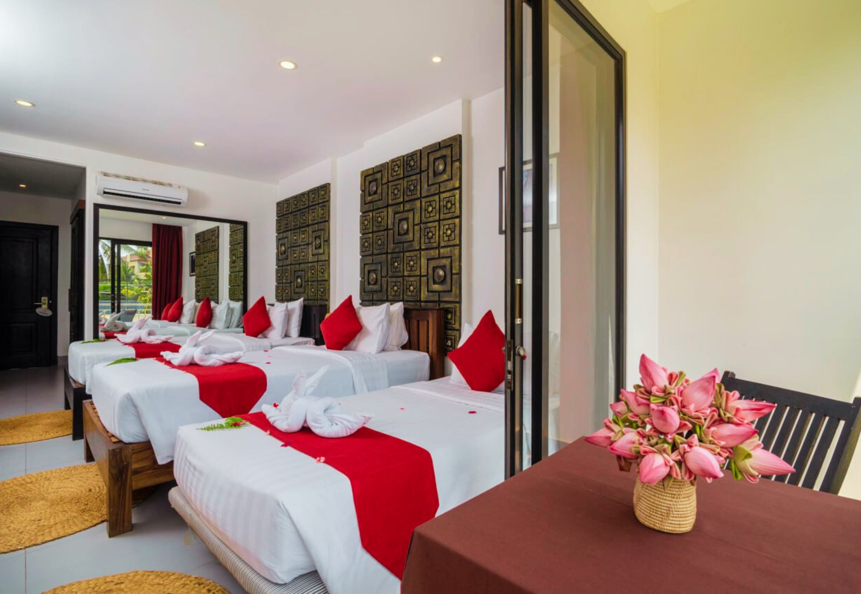 A hotel room with three beds, each with white linens and red accents, a wooden floor, and a view of greenery outside.