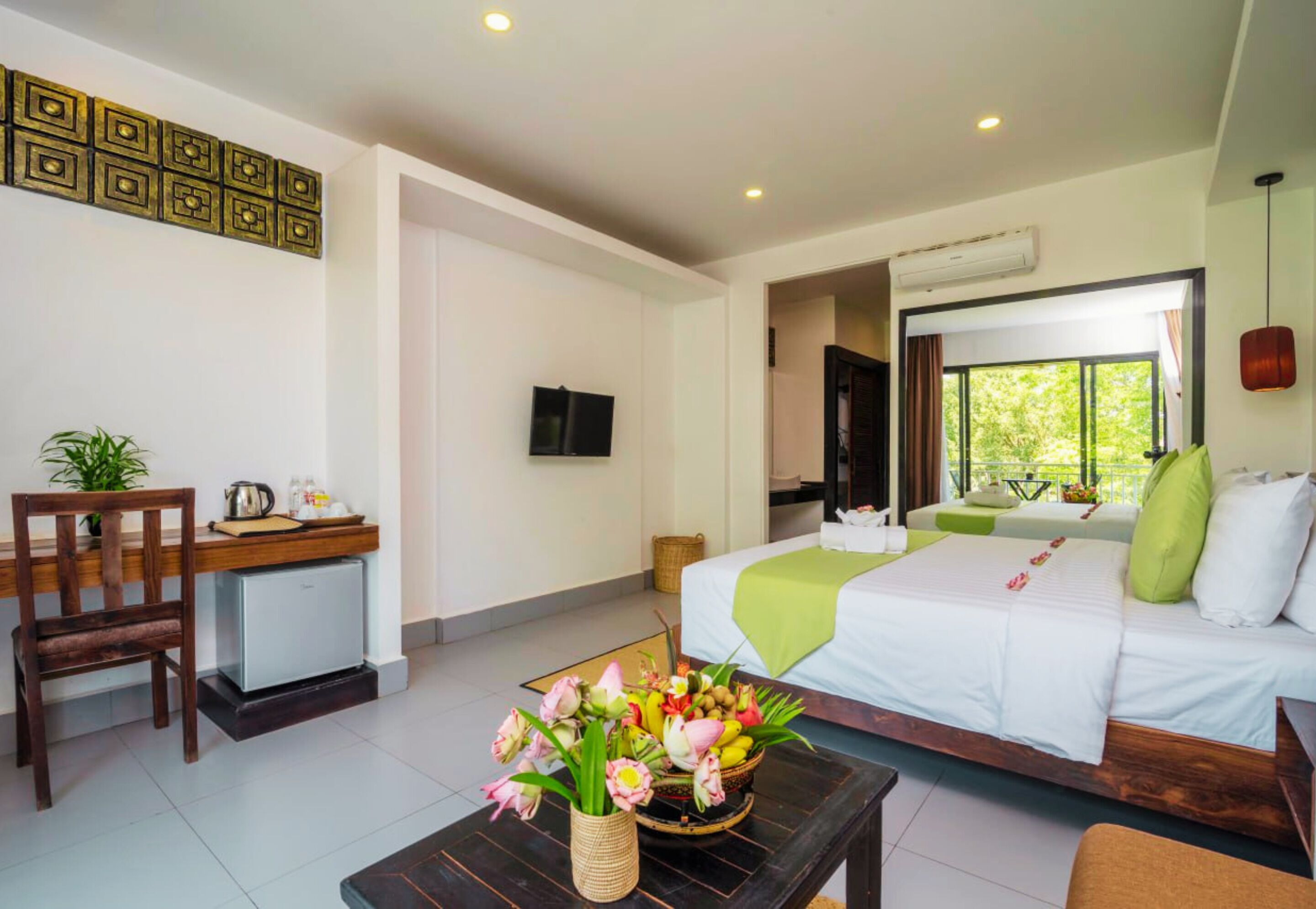 A hotel room with a bed, a desk with a chair, a small refrigerator, a television, and a window with a view of greenery.