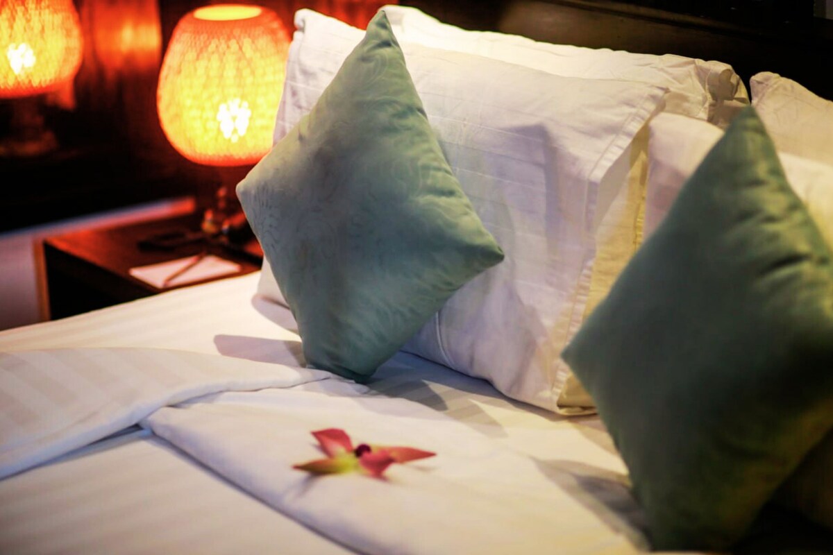 A neatly made bed with white linens and green pillows, a red flower decoration, and a lamp in the background.