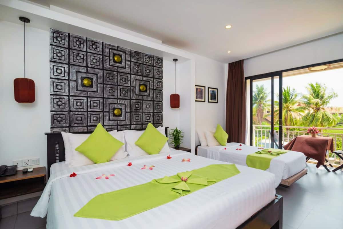 Habitación de hotel con dos camas, una pared decorativa, un balcón con vistas a palmeras y una pequeña mesa con una silla.