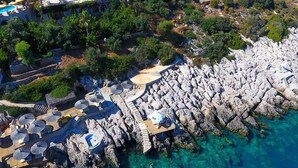 An aerial view of a coastal hotel with a rocky shoreline, clear blue waters, and a deck area with umbrellas. - Hotel Marsala (Kas)