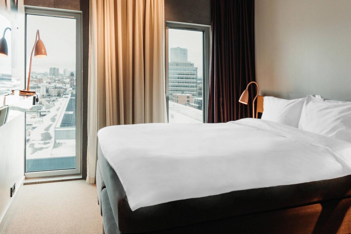 Una camera d'albergo con un letto, una finestra con tende e vista sulla città, e una lampada.