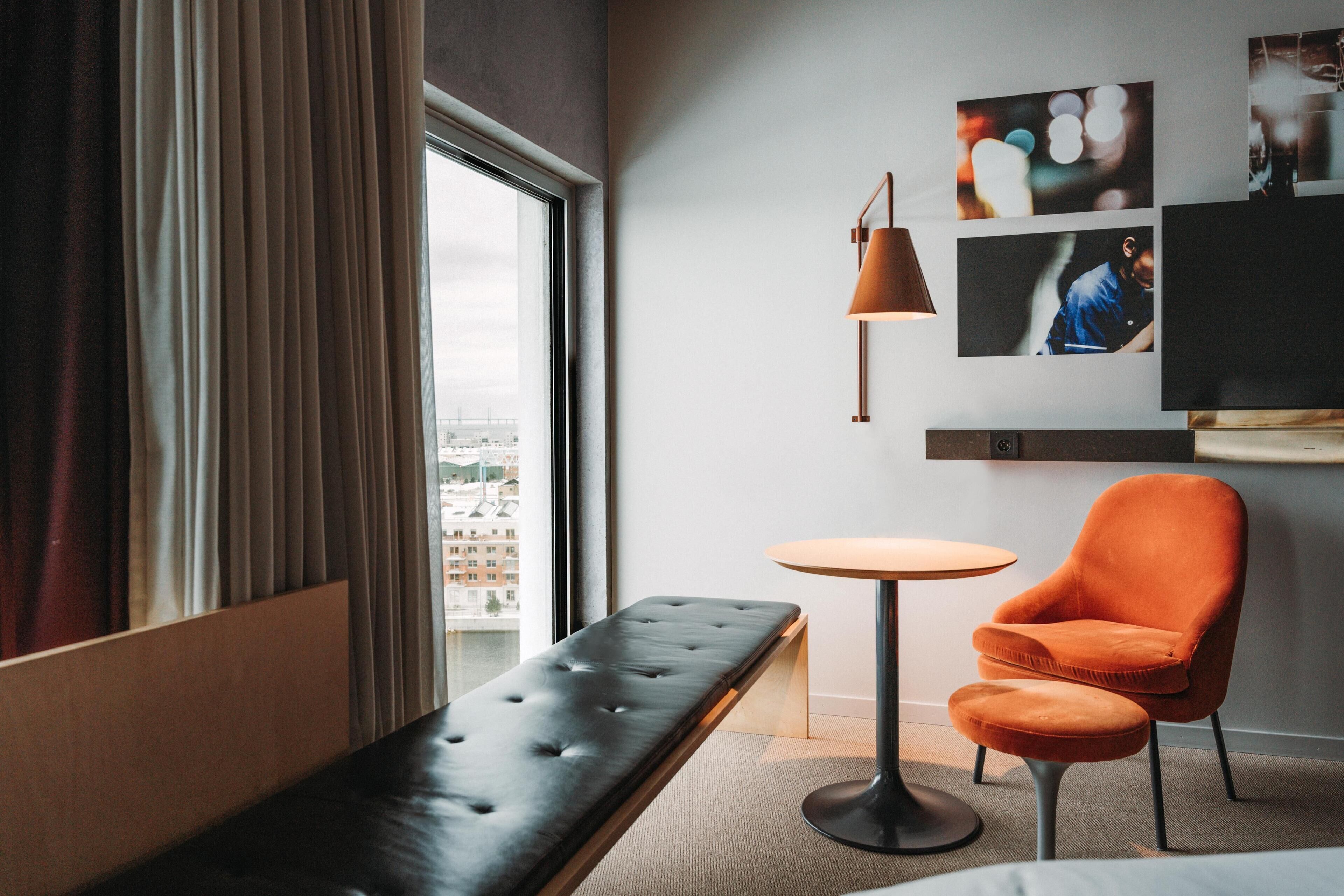 Una habitación de hotel con un ventanal grande, un banco, una silla naranja, una mesa redonda y un televisor montado en la pared.