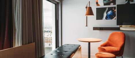 Una habitación de hotel con un ventanal grande, un banco, una silla naranja, una mesa redonda y un televisor montado en la pared.
