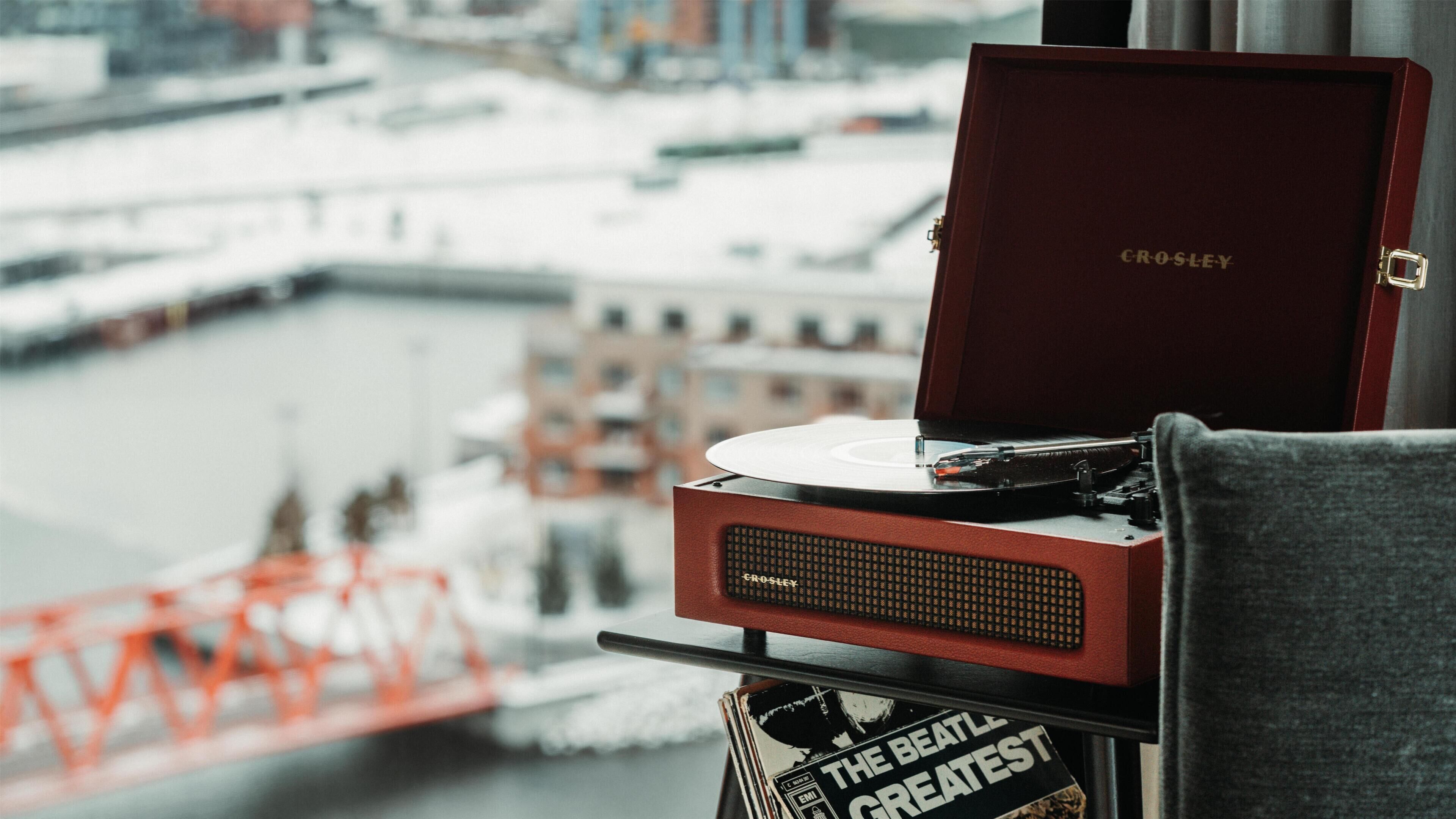 Un tocadiscos Crosley sobre una mesa con un disco de vinilo, un libro titulado 'The Beatles Greatest Hits', y una vista de un río con edificios y un puente al fondo.
