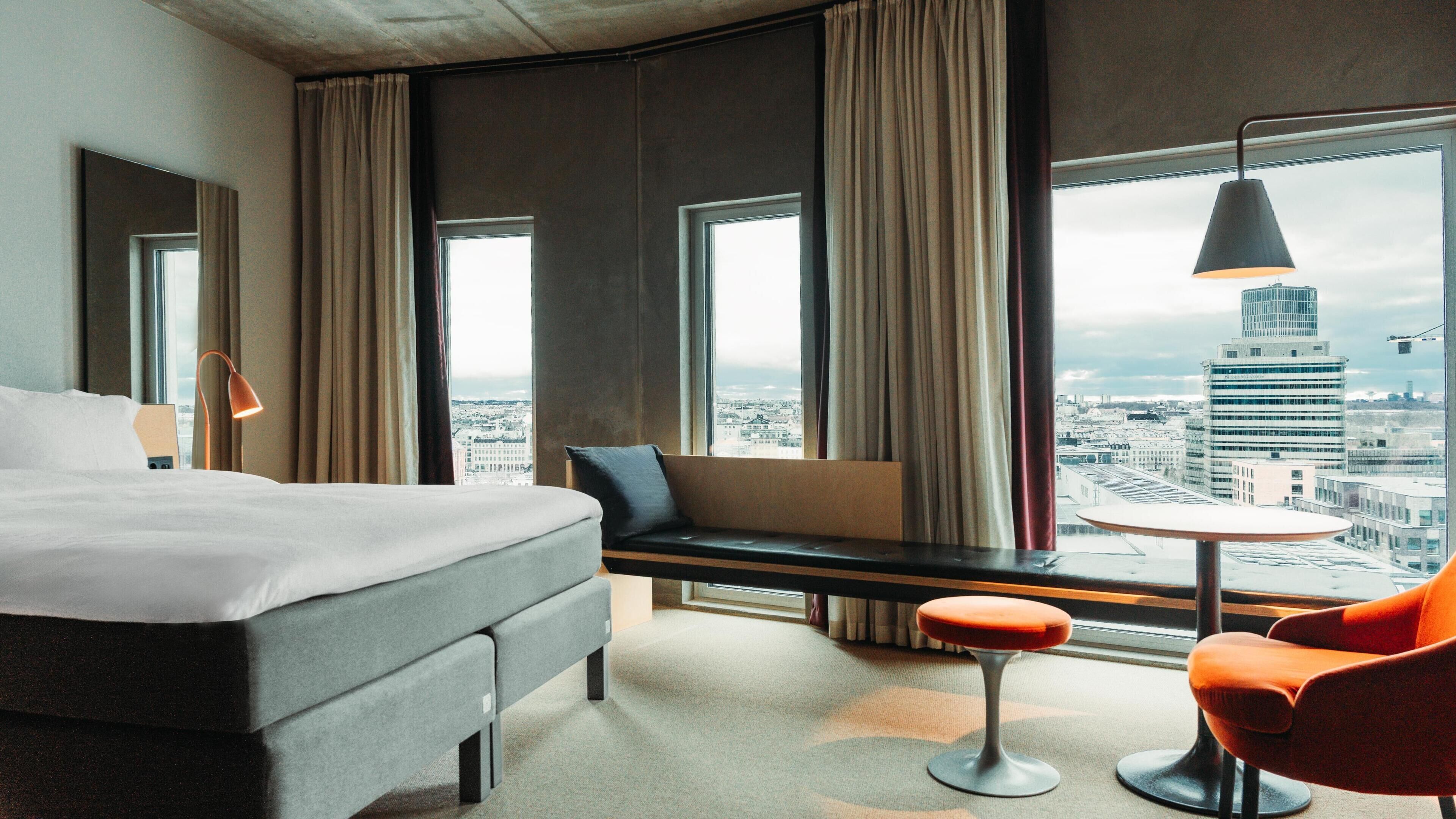 A hotel room with a bed, a sofa, a round table, and a chair, with a city view through large windows.