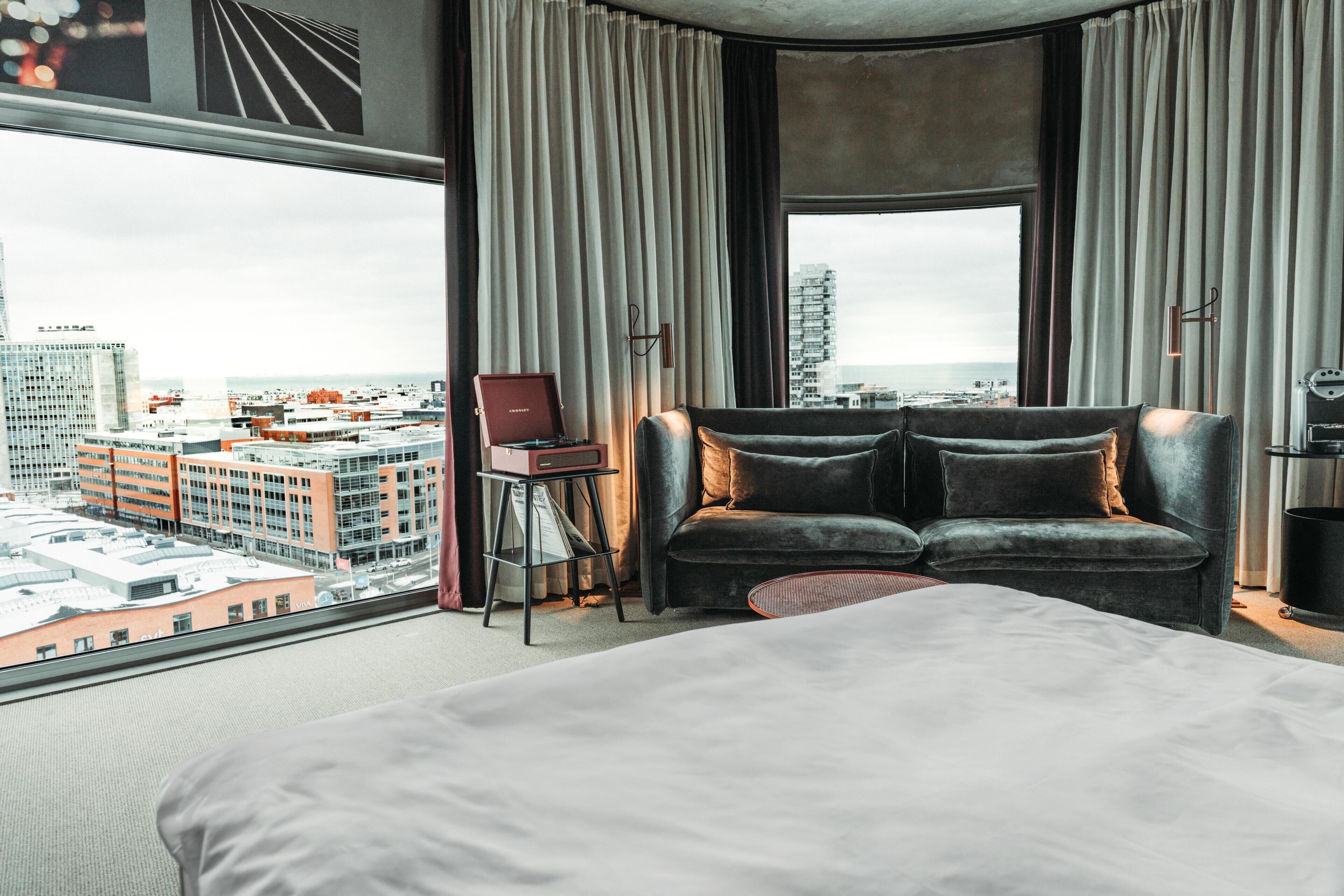 A hotel room with a large bed, a sofa, a chair, and a view of the cityscape.