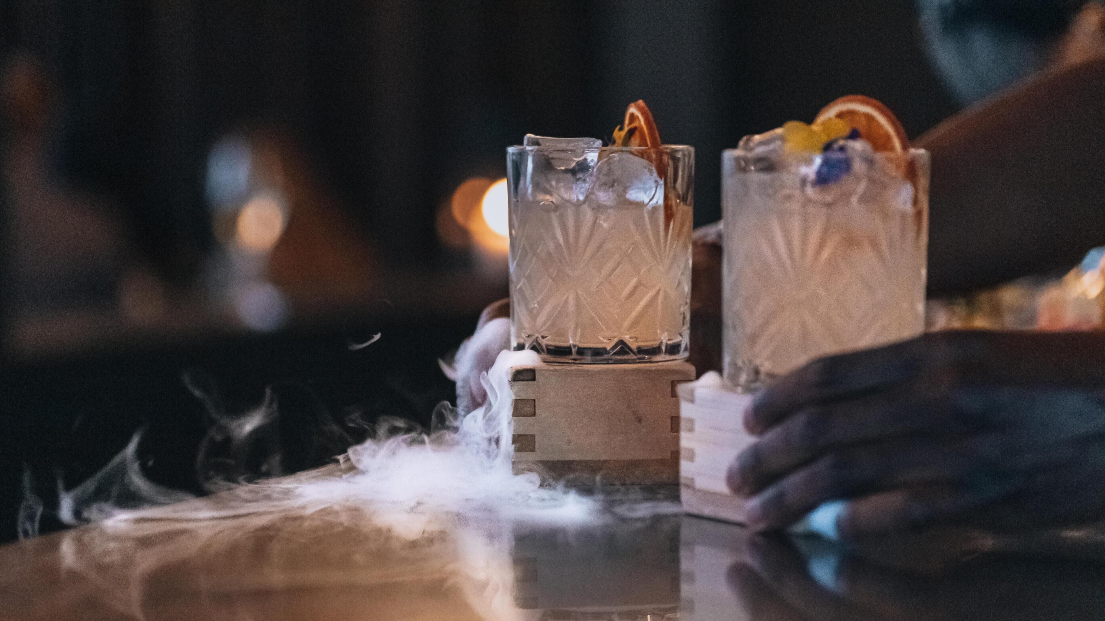 Two glasses of a clear beverage with ice and garnishes on a bar counter.