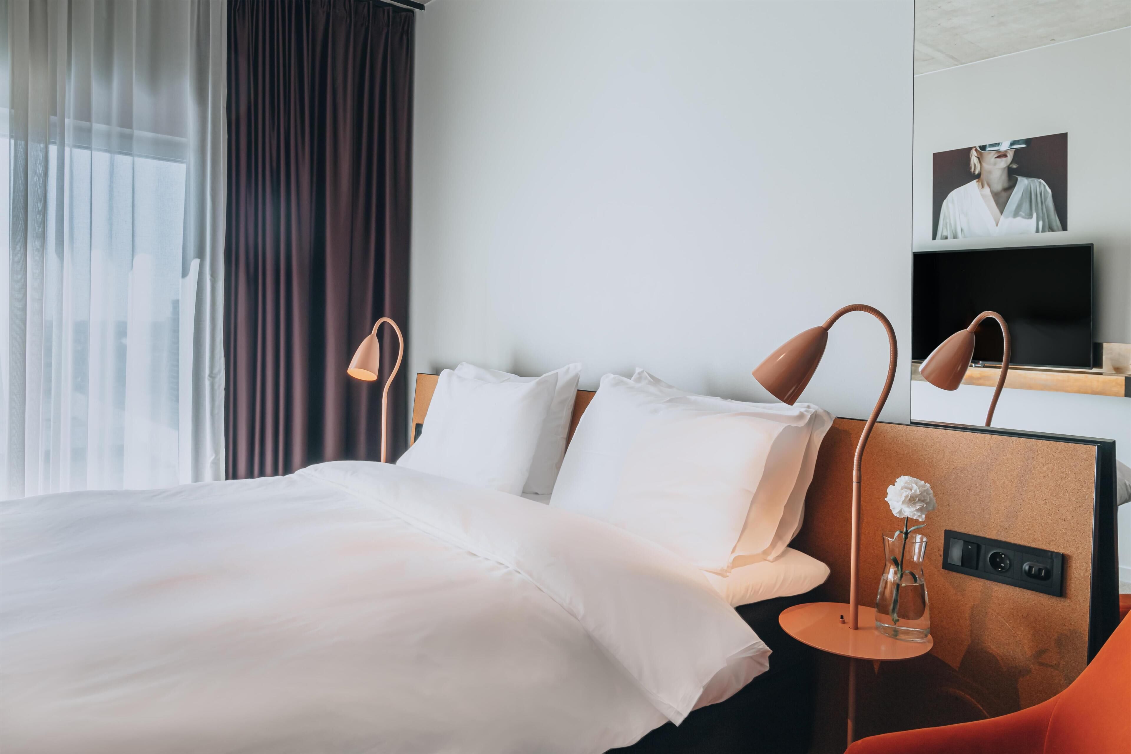 A hotel room with a bed, a TV, a vase with a flower, and a window with curtains.