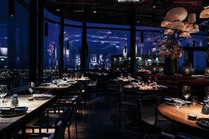 Um restaurante com mesas postas para refeições, amplas janelas com vista para a cidade e lanternas de papel penduradas.