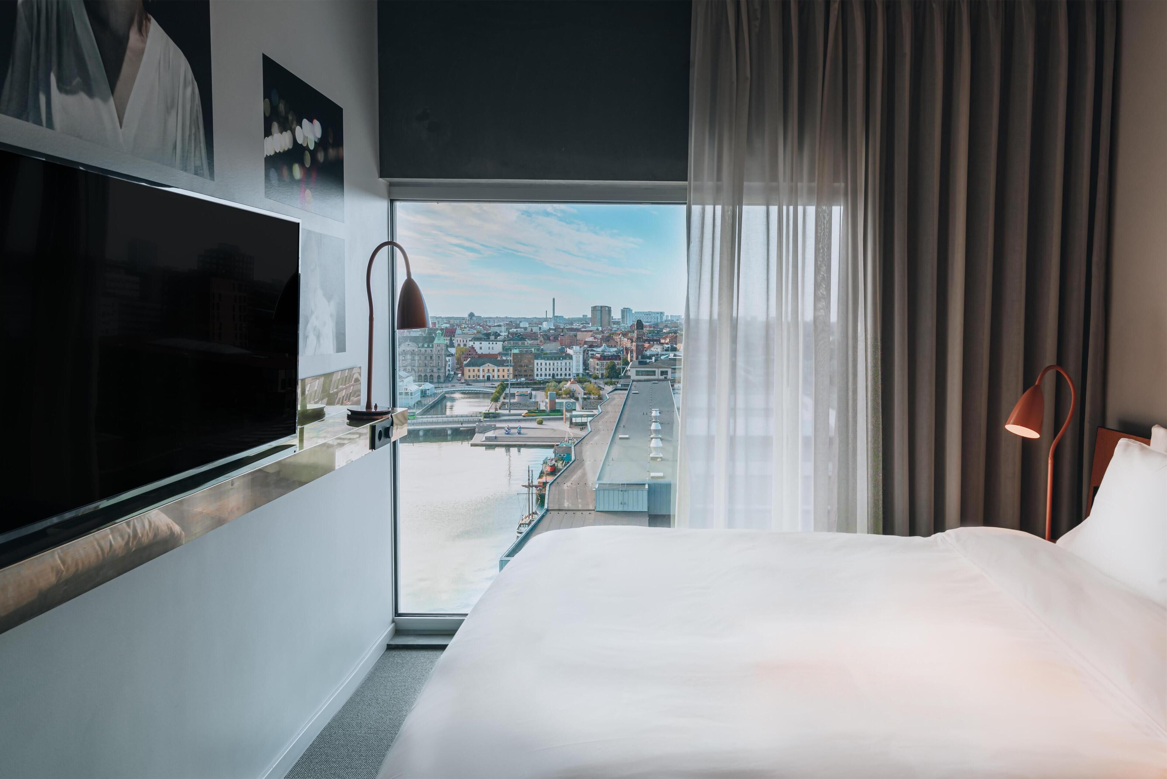 Et hotelværelse med en stor seng, fjernsyn, udsigt over byen og et vindue med gardiner.