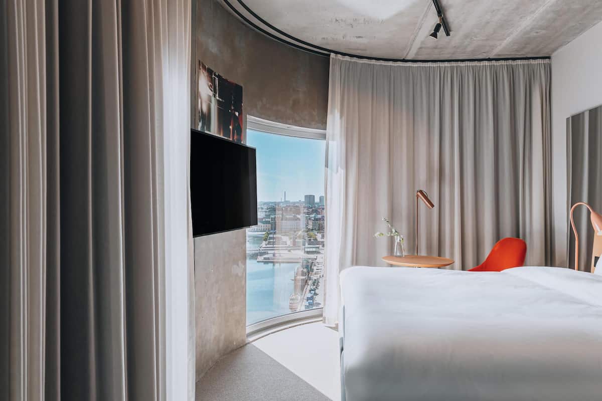 Una camera d'albergo moderna con un letto grande, una TV a parete, vista sulla città dalla finestra e una poltrona rossa.