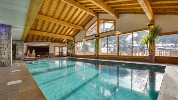 An indoor swimming pool with clear blue water, wooden beams, stone pillars, and large windows offering a view of snow-covered houses. - CGH Residence Boutique les Chalets de Léana (Araches-la-Frasse)