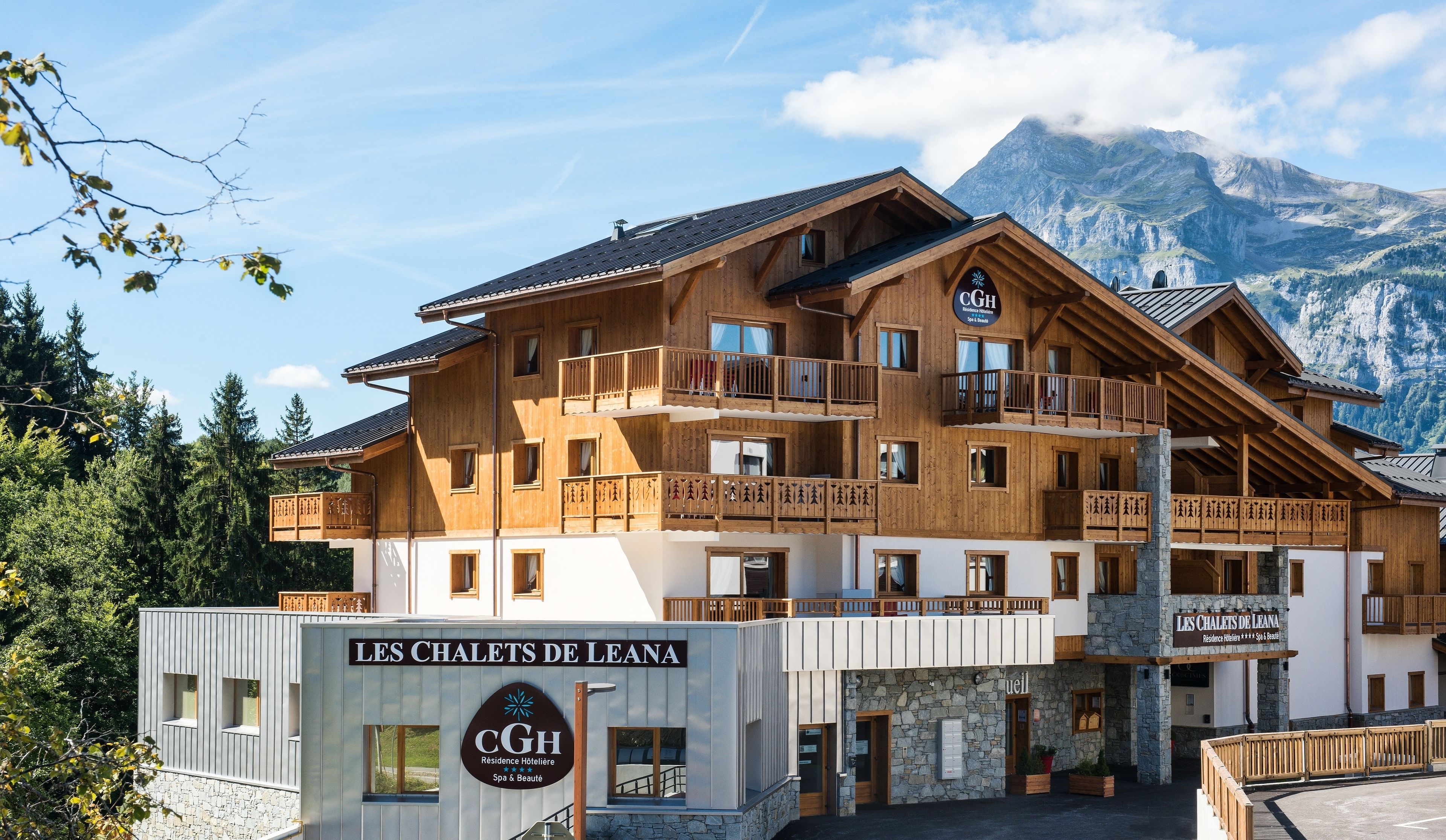 A multi-story wooden building with balconies, a sign for 'Les Chaquets de Leana', and a mountain backdrop.