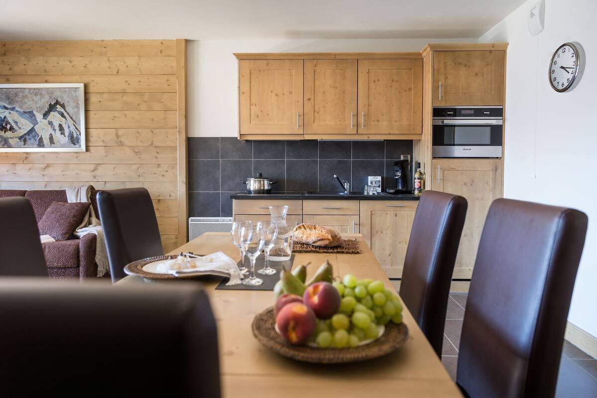 A dining area with a wooden table set for four, a fruit bowl, and a clock on the wall.