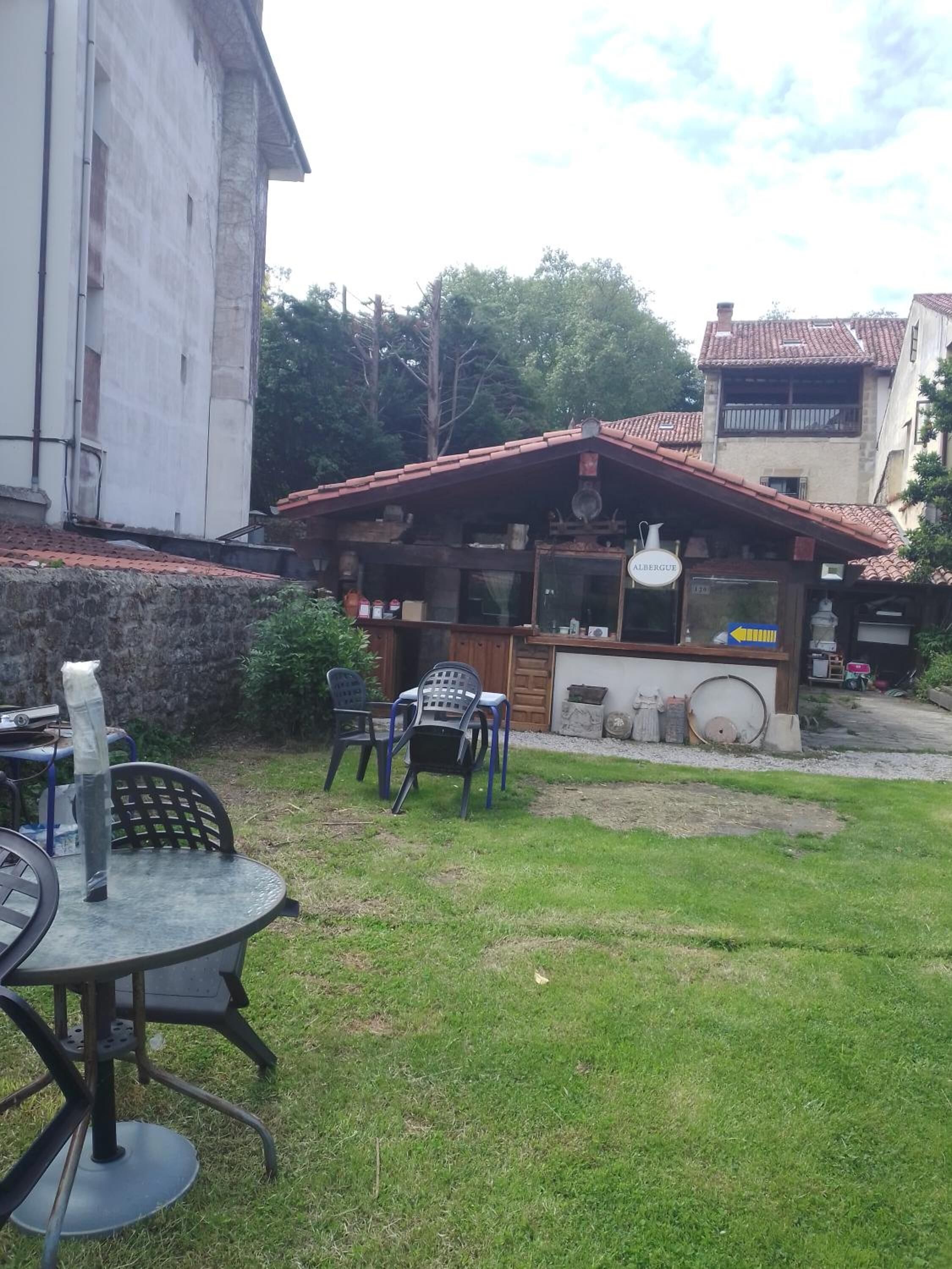 A small outdoor seating area with a table and chairs, a wooden structure with a tiled roof, and a building in the background.