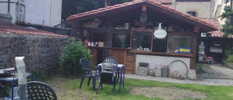 A small outdoor seating area with a table and chairs, a wooden structure with a tiled roof, and a building in the background.