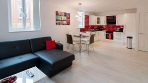 Een moderne woonkamer met een zwarte bank en een eettafel met stoelen. Er is een keukenhoek met rode tegels en witte kastjes.