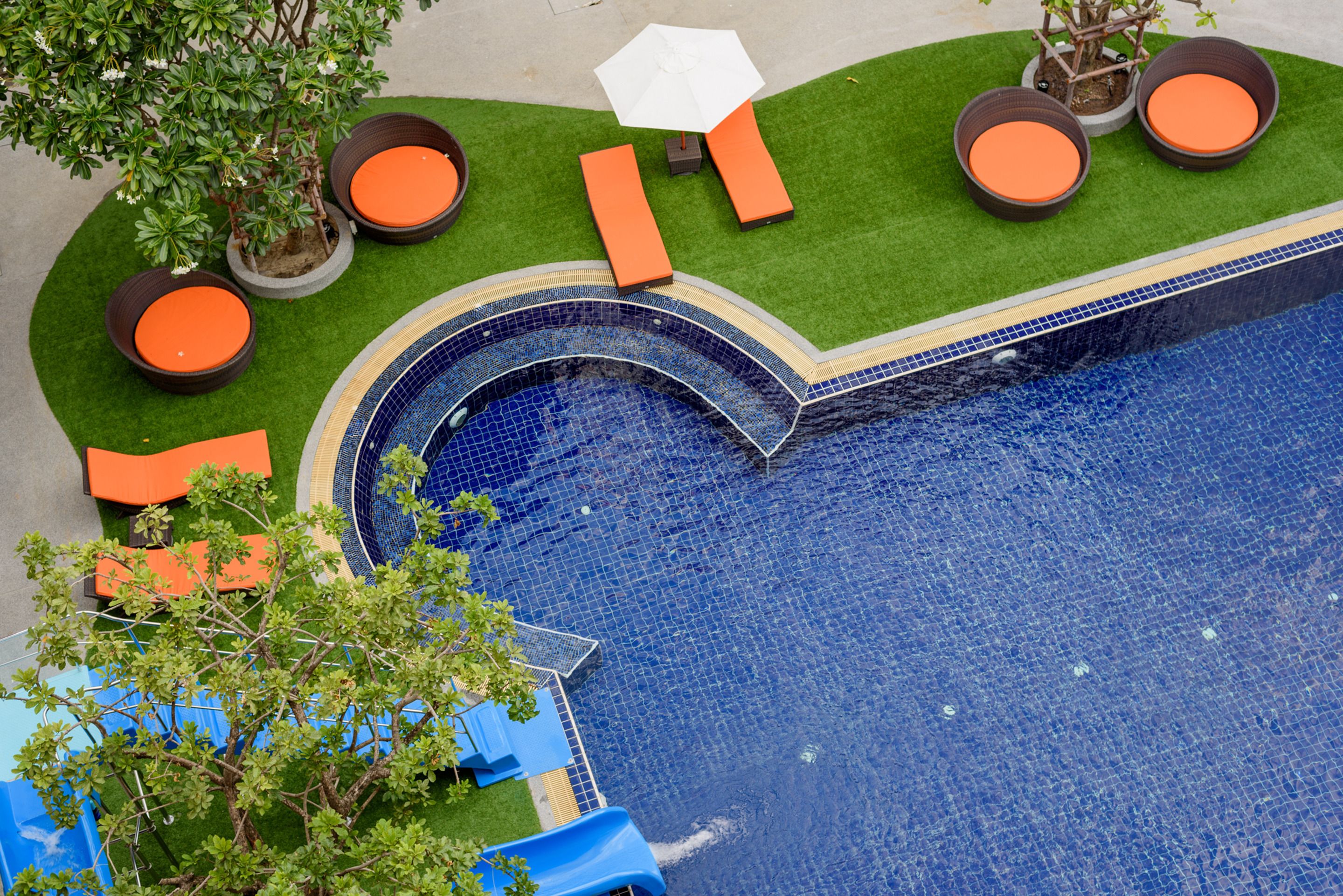 Piscina com borda curva, rodeada por grama sintética e assentos laranja.