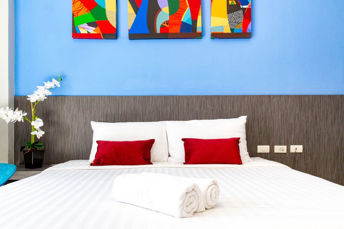 Una cama bien hecha con almohadas blancas y rojas, una toalla blanca enrollada y un florero con flores blancas sobre una mesita de noche de madera.