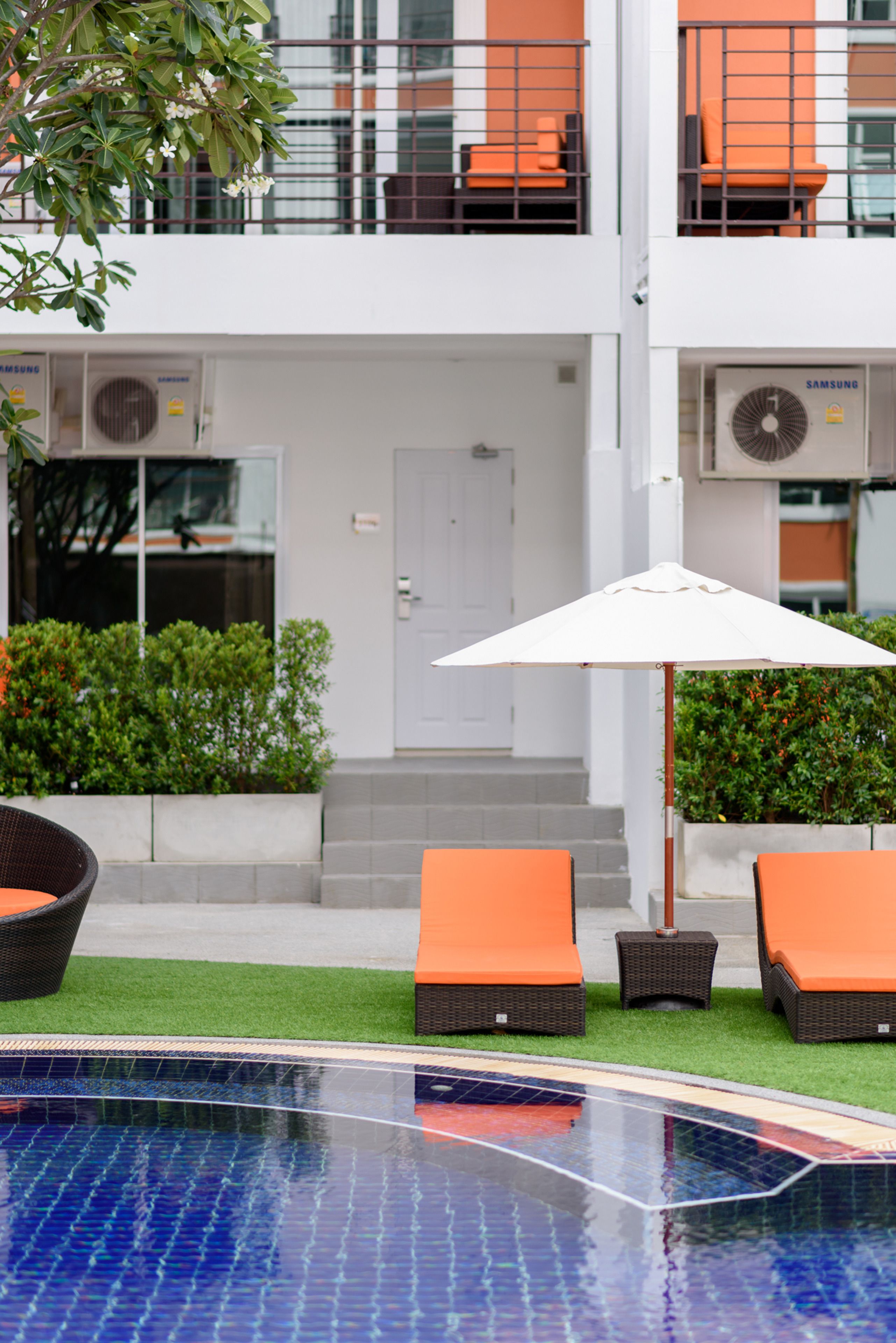 Zona de piscina con sillones de descanso y una sombrilla, rodeada por un edificio con unidades de aire acondicionado y una puerta.