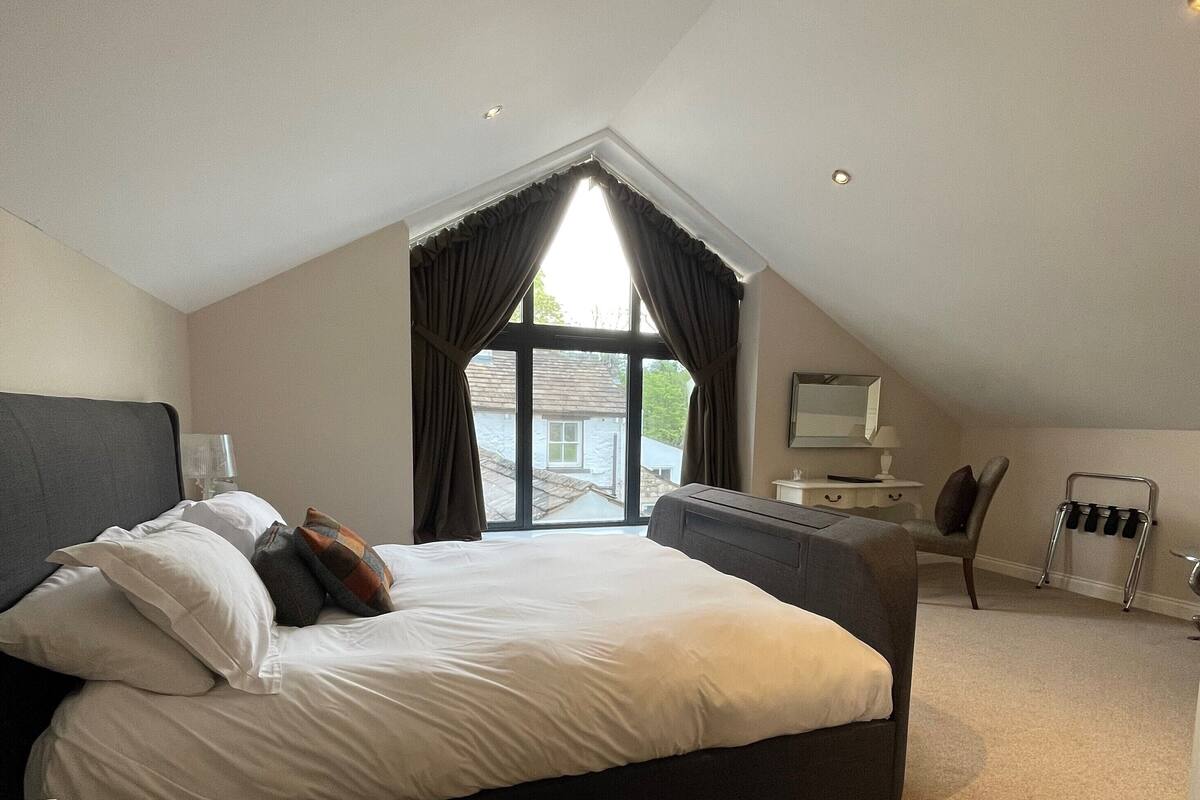A bedroom with a large bed, a desk, a chair, and a window with curtains.