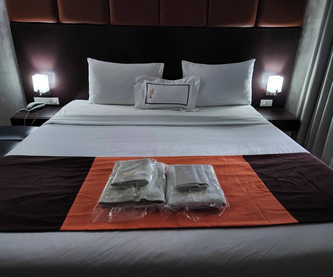 A neatly made bed with white linens, a pillow with a red logo, and two rolled towels on an orange mat.