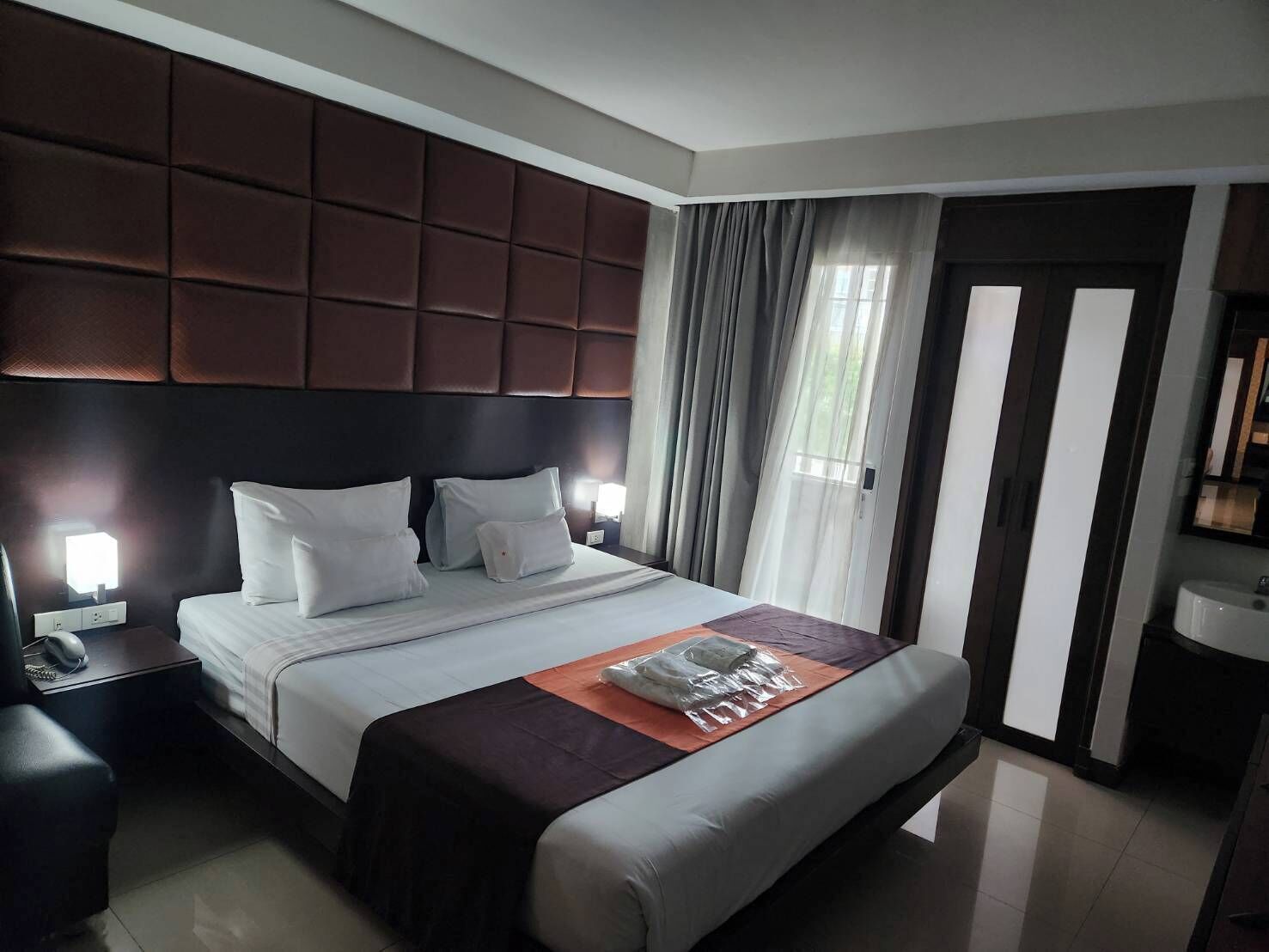 Ein modernes Hotelzimmer mit einem großen Bett, einer gepolsterten Kopfteil, einem Nachttisch mit Lampe und Blick durch Vorhänge nach draußen.