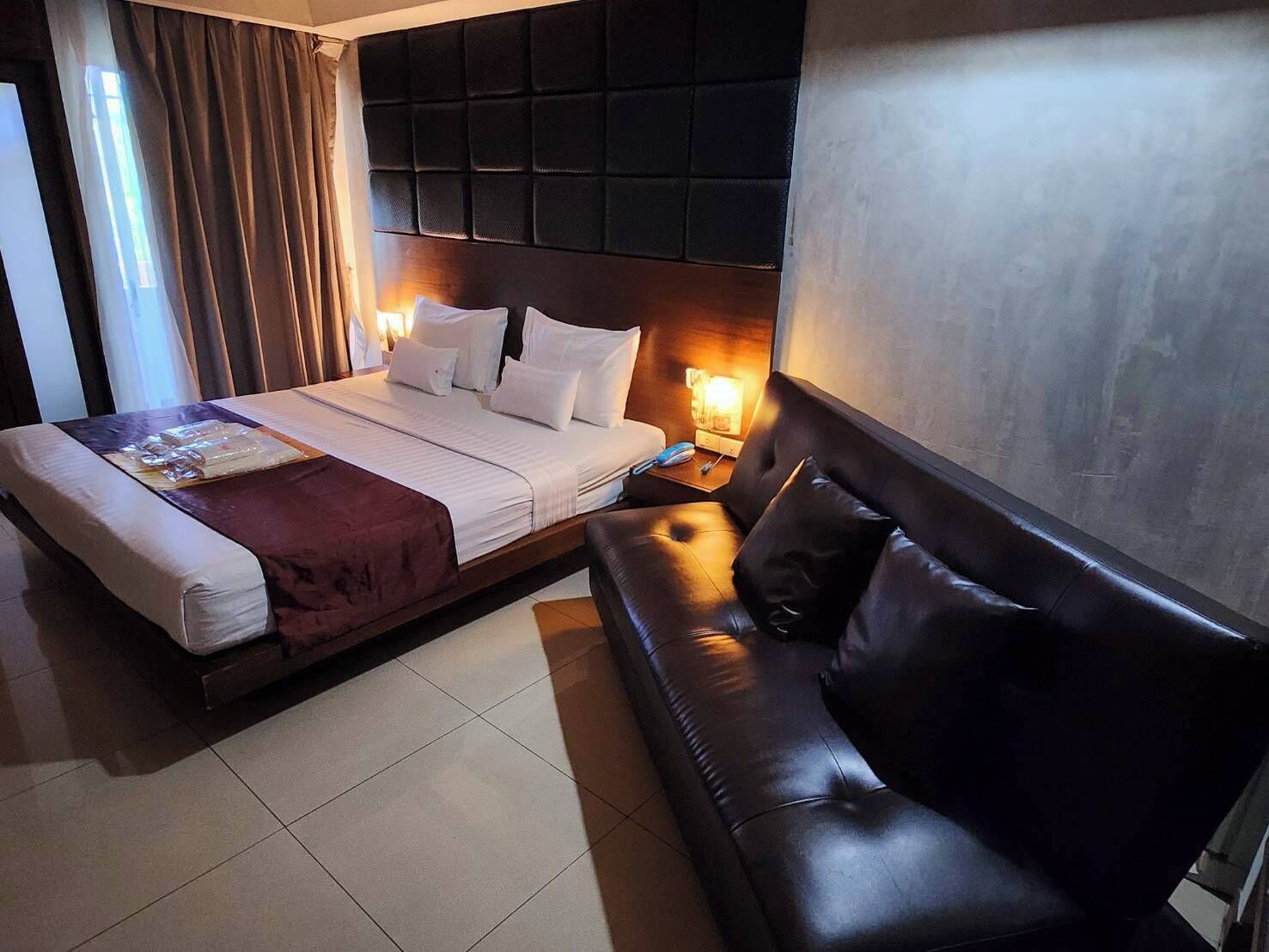 Ein Hotelzimmer mit Bett, dunklem Kopfteil, Nachttisch mit Lampe und einer Ledersofa.